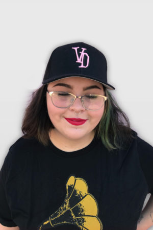 Woman wearing a black voodoo doughnut flexfit hat with pick VD initials